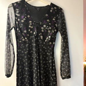 Svart blommig klänning - Svart kortklänning med små vita och lila blommor. Klänningen har transparenta långärmade ärmar, v-ringning och knyt framtill. Tunn mesh-liknande polyester. 100% polyester