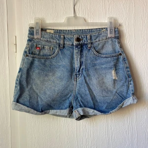 Blå jeansshorts med upprullad kant - Klassiska blå jeansshorts från Levi's med upprullad kant och femficksmodell. Shorts med normal passform, hög midja och en liten sliten detalj framtill. Perfekta för sommaren!