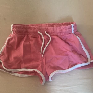 Rosa sportiga shorts med vit kant - Säljer ett par rosa sportiga shorts med vit kant och snörning i midjan. De har elastisk midja och är perfekta för träning eller varma sommardagar. Mjukt bomullsmaterial som känns skönt mot huden.