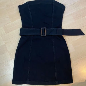 Svart axelbandslös klänning med bälte - Säljer en svart axelbandslös klänning med markerad midja och brett bälte med stor spänne. Klänningen har en figurnära passform och dragkedja bak. Perfekt för dig som vill ha en stilren och enkel look.