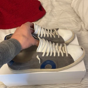 Grå sneakers med blå detalj från Oliver Cabell - Säljer ett par grå sneakers från Oliver cabell med vit sula, vita skosnören och blå hälkappa. Skorna har en rund tå och en blå cirkeldetalj på sidan. Perfekta för en avslappnad stil och passar till många outfits.