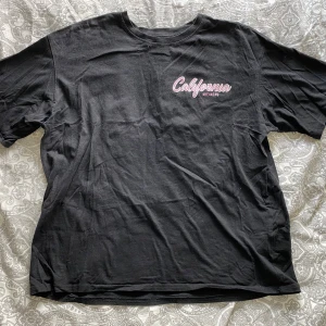 Svart t-shirt med tryck från SHEIN - Svart t-shirt från SHEIN med tryck i rosa och vitt. På bröstet står det 'California' och på ryggen finns ett större tryck med palmer och texten 'Summer Adventures, West Coast Surf Riders'. Klassisk passform och rund hals.