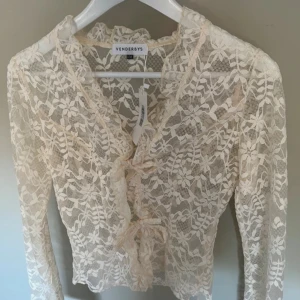 Vit spetsblus från Venderbys - Säljer en elegant vit spetsblus från Venderbys med långa ärmar och v-ringning. Blusen har fina volangdetaljer framtill och knytning vid bysten. Perfekt för dig som gillar romantisk stil och transparenta material.