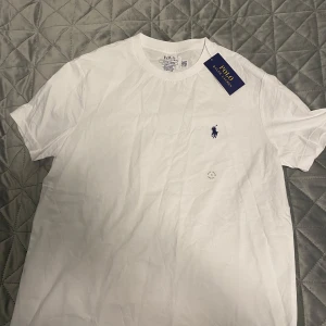 Vit t-shirt från Polo Ralph Lauren - HELT OANVÄND. Klassisk vit t-shirt från Polo Ralph Lauren med blå broderad logga på bröstet. Modellen är slim fit och har rund hals och korta ärmar.