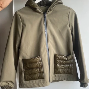 Olivgrön jacka med luva från JOTT - Snygg olivgrön jacka från JOTT med glansiga quiltade detaljer på fickor och huva. Jackan har dragkedja framtill, två fickor och en mjuk insida (fleece). Hittar ingen storleksmärkning men köpt på ”damavdelningen” second hand & skulle gissa att det är en stl XS