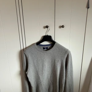Gant tröja - Säljer en klassisk grå stickad tröja från GANT i storlek S. Tröjan har rund halsringning med blå och vit randig detalj på insidan av kragen. Perfekt för lager-på-lager och enkel att matcha.