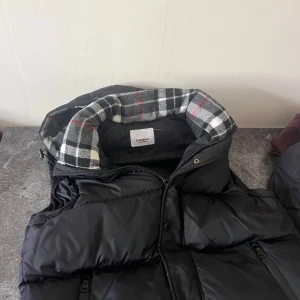 Svart dunväst från Burberry - Säljer en svart dunväst från Burberry med stor logotyp på bröstet. Västen har dragkedja och en fodrad huva med rutigt mönster i svart, vitt och rött. Perfekt för kyliga dagar när du vill ha både stil och värme.