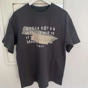 Svart t-shirt från Maison Margiela - Nu säljer vi denna svarta t-shirt från Maison Margiela med tryck av siffror och logga på bröstet. Klassisk rund hals och korta ärmar. Perfekt för dig som gillar streetwear och designerplagg.