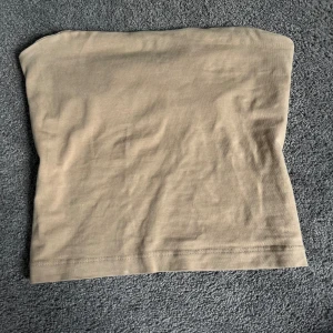 Beige tubtopp från Gina Tricot - Säljer en enkel och stilren beige tubtopp från Gina Tricot. Toppen är ärmlös och har en rak passform, perfekt att matcha med jeans eller kjol. Tillverkad i mjuk bomull för en bekväm känsla.