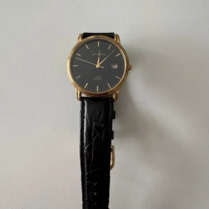 Svart armbandsur från Leijona med guldfärgad boett - Klassisk klocka från Leijona med svart urtavla, guldfärgad rund boett och datumvisning. Svart läderarmband med krokodilpräglad yta. Passar dig som gillar stilrena accessoarer. Endast bandet behöver bytas men själva uret för perfekt.