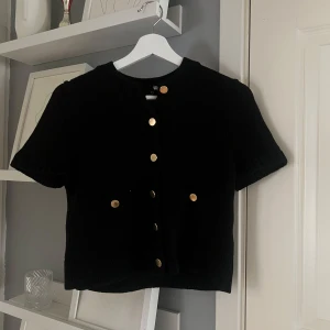 Svart stickad kofta med guldknappar - Kortärmad svart stickad kofta med rund hals och dekorativa guldknappar framtill. Två små fickor framtill. Perfekt att styla över en topp eller klänning.