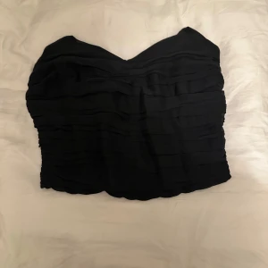 Svart croppad tubtopp - Säljer en svart croppad tubtopp med volangliknande detaljer i flera lager. Toppen har en hjärtformad urringning och är perfekt för att skapa en trendig look.