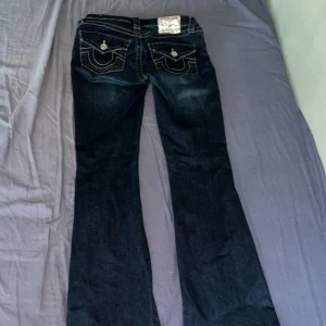 Mörkblå bootcut jeans från True Religion - Säljer ett par mörkblå jeans från True Religion med bootcut passform. Jeansen har kontrastsömmar, klassiska fickor med lock och knapp bak samt tydlig logga på bakfickan. Perfekta för dig som gillar en tidlös jeansstil. (Säljer för att dem är förstora för mig)