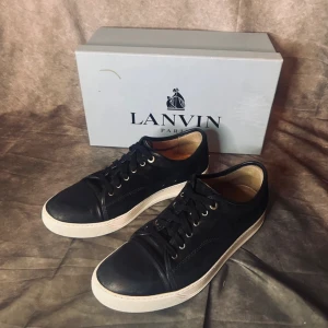 Blåa sneakers från Lanvin - Säljer ett par svarta sneakers från Lanvin med vit platt sula och rund tå. Skorna har snörning och är tillverkade i skinn med diskret logga på sulan. Klassisk och stilren design som passar till mycket. Tar byten