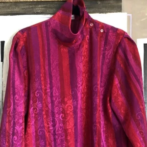 Röd/ros  och lila mönstrad långärmad blus från Kurt Kellermann - långärmad blus från Kurt Kellermann i röd/rosa och lila med breda ränder och diskret blommönster. Hög krage med knappar som kan bäras på olika sätt. Perfekt för dig som vill sticka ut med färg och retrostil. Ingen markerad strl men skulle säga den passar bäst 40/42