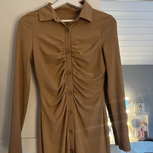 Beige långärmad klänning med knappar och rynk - Säljer en beige långärmad klänning med krage och knappar hela vägen framtill. Klänningen har snygga rynkdetaljer längs knappraden och är figurnära i modellen. Perfekt för dig som gillar stilrena och trendiga plagg.