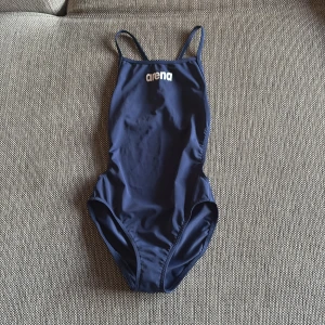 Mörkblå baddräkt från Arena - En stilren mörkblå baddräkt från Arena med smala axelband och klassisk sportig design. Perfekt för simträning eller bad. Logotypen är tryckt framtill i vitt.