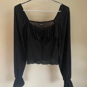 Svart blus med rynkade detaljer - Svart blus från Gina Tricot med rynkade detaljer och fyrkantig ringning. Långa ärmar med resår vid handlederna och lätt croppad passform. Perfekt att styla med jeans eller kjol.