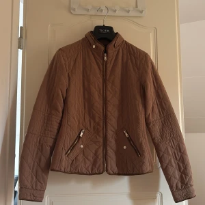 Quiltad brun jacka från lft  - Snygg quiltad brun jacka från lgt med dragkedja framtill och två fickor med dragkedja. Jackan har hög krage och långärmade ärmar. Perfekt för kyligare dagar och ger en stilren look.