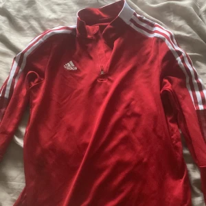 Röd långärmad träningströja från Adidas - Säljer en röd långärmad träningströja från Adidas med vita ränder längs ärmarna och dragkedja vid halsen. Tröjan har klassisk Adidas-logga på bröstet och är tillverkad i ett lätt syntetmaterial.