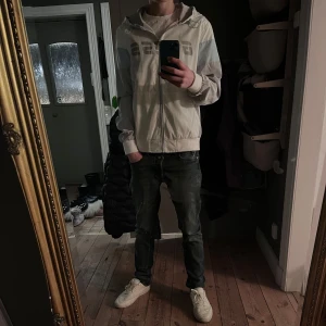 Helt ny vindjacka från Guess - Säljer en stilren vit och ljusgrå vindjacka från Guess med huva och dragkedja. Använd endast 1 gång. Jackan har stora fickor framtill och Guess-logga på bröstet. Vattenavvisande material, perfekt för vår/sommar/höst. NYPRIS: 1500kr