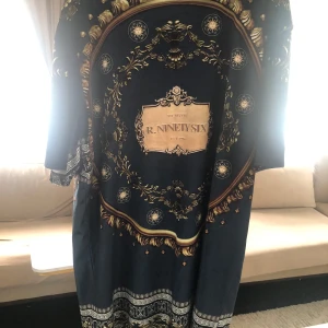 Mörkblå mönstrad kimono från R. Ninety Six - Säljer en mörkblå kimono med lyxigt barockinspirerat mönster i guld och vitt från R. Ninety Six. T-shirts har vida ärmar och ett iögonfallande tryck på ryggen. Perfekt för dig som vill sticka ut med en unik och elegant stil.