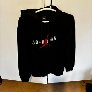 Svart hoodie med tryck från Joarlean - Svart hoodie med vit text och röd figur på bröstet. Klassisk huva med dragsko och magficka framtill. Perfekt för en avslappnad stil. stlk XS/S. Använd 1 gång!