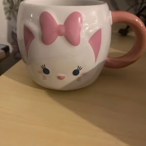Söt kattmugg med rosa rosett - En vit keramikmugg med ett kattansikte, rosa öron och en stor rosa rosett framtill. Handtaget är också rosa. Perfekt för te eller kaffe och har en gullig design som passar kattälskare.
