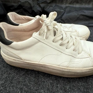 Vita sneakers med svart häl - Säljer ett par vita sneakers med svart detalj på hälen. Skorna har snörning och en platt sula. Perfekta för en avslappnad och stilren look. Passar till det mesta och är riktigt bekväma att gå i.