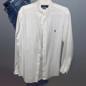 Vit skjorta från Polo Ralph Lauren - Klassisk vit skjorta från Polo Ralph Lauren i slim fit-modell. Skjortan har lång ärm, knappar framtill och en broderad logga i blått på bröstet. Perfekt för en stilren och tidlös look.