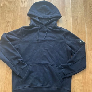 Svart hoodie från Calvin Klein Jeans - Säljer en svart hoodie från Calvin Klein Jeans med stor CK-logga på ärmen. Tröjan har huva med dragsko och en stor magficka framtill. Perfekt för dig som gillar stilrena och klassiska plagg.