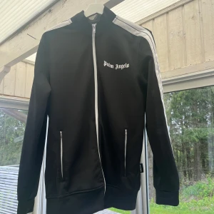 Svart track jacket från Palm Angels - Svart track jacket från Palm Angels med vit dragkedja och vita ränder längs ärmarna. Jackan har hög krage, två fickor med dragkedja och logga på bröstet. Perfekt för en sportig och trendig look.