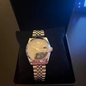 Seiko mod datejust  - Stilren egenbygd Seiko mod klocka i silver med grå/silver urtavla med romerska siffror. Det är en 40mm datjust. Klockan har datumvisning och länkarmband i rostfritt stål (jubilee armband). Perfekt nu inför sommaren! Skriv järna vid frågor och funderingar!