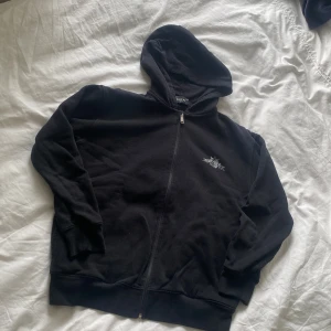Svart hoodie med dragkedja från Replay - Svart hoodie från Replay med dragkedja framtill och huva. Liten logga på bröstet och stor REPLAY-text baktill. Mjuk insida och ribbade muddar. Perfekt för en avslappnad stil.