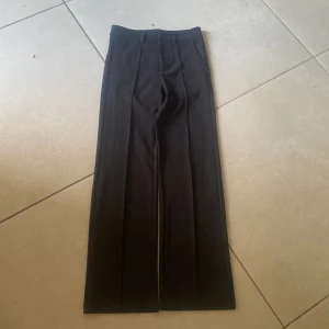 Svarta kostymbyxor med pressveck - Säljer ett par svarta kostymbyxor med raka ben och pressveck framtill. Byxorna har hög midja och klassisk passform, perfekta till en stilren look. Materialet känns mjukt och följsamt, och de är utan synliga detaljer som knappar eller dragkedja på framsidan.