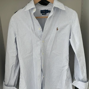 Blårandig skjorta från Ralph Lauren - Säljer en klassisk blårandig skjorta från Ralph Lauren i modellen Custom Fit. Skjortan har långa ärmar, knappar framtill och en broderad logga på bröstet. Perfekt för en stilren och fräsch look! Kan tyvärr ej lösa bild med den på då den är för liten för mig