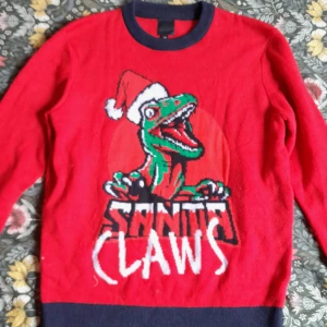 Röd jultröja med dinosaurie från H&M - Rolig röd jultröja från H&M med en grön dinosaurie som har tomteluva och texten 'Santa Claws'. Mörkblå muddar vid hals, ärmslut och nederkant. Perfekt för julens alla tillfällen!