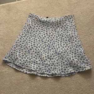 Vit kjol med blått blommönster - Supersöt vit kjol med små blå blommor över hela tyget. Kjolen har en lätt utställd form och är perfekt för varma dagar. Den är kort och har en mjuk, flowy känsla. Perfekt att styla med en enkel topp eller hoodie.