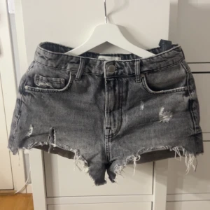 Grå jeansshorts med slitningar - Säljer ett par grå jeansshorts från zara med råa kanter och slitna detaljer framtill. Klassisk femficksmodell med knapp och dragkedja. 