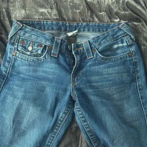 Blå jeans från True Religion - Säljer ett par klassiska blå jeans från True Religion med kontrastsömmar och ikonisk fickdesign. Jeansen har bootcut passform och är tillverkade i denim. Storlek 28, tillverkade i USA.