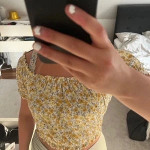 Blommig offshoulder topp med puffärmar - Söt offshoulder topp med gula och grå blommor på vit botten. Puffiga korta ärmar och resår vid axlar och midja för en smickrande passform. Perfekt för varma dagar och somriga outfits.