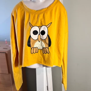 Gul croppad tröja med uggletryck - Säljer en gul croppad tröja med ett stort, tecknat uggletryck på framsidan. Tröjan har långa ärmar och en avslappnad passform med bred halsringning. Perfekt för dig som gillar färg och roliga motiv!