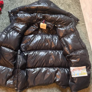 Moncler montbeliard  - Säljer en svart, glansig dunjacka från Moncler med klassisk logotyp på bröstet och rödvit dragkedja. Jackan har hög krage och stora, vadderade sektioner som ger en puffer-look. Perfekt för kalla dagar.