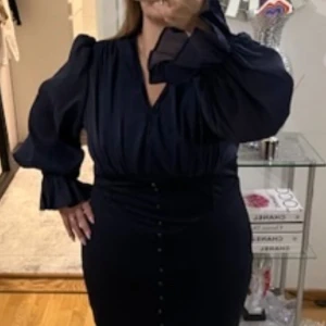 Marinblå långklänning med knappar och v-ringning - Elegant marinblå långklänning med v-ringad hals, långa puffärmar och dekorativa knappar framtill. Klänningen har markerad midja och lätt glansigt tyg som ger en lyxig känsla. Perfekt för speciella tillfällen.