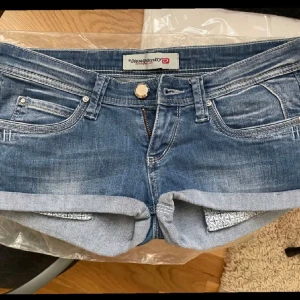 Blå jeansshorts från Diesel Industry - Säljer ett par klassiska blå jeansshorts från Diesel Industry med uppvikta benslut och femficksmodell. Shortsen har låg midja och snygga slitningar för en avslappnad look. Perfekta för sommaren!