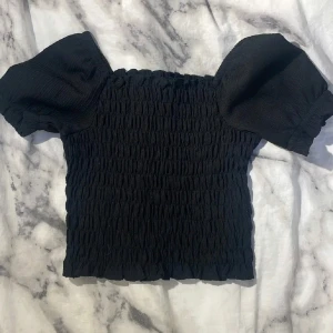 Svart topp med puffärmar från WE - Säljer en svart topp från WE med korta puffärmar. Toppen har en elastisk, rynkad struktur och är perfekt för dig som gillar trendiga detaljer. Passar till många olika stilar.