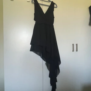 Svart asymmetrisk klänning med volang och v-ringning - Säljer en svart klänning med djup v-ringning och asymmetrisk volangkjol. Klänningen har omlottkänsla och knyt på bak sidan, vilket ger en snygg siluett. Perfekt för dig som vill sticka ut med en elegant och modern look.Helt ny.Storlek passar både M och L