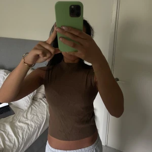 Brun ribbad ärmlös topp - Säljer en brun, ribbad och croppad ärmlös topp med hög hals. Perfekt för dig som gillar en enkel men stilren look. Toppen har en tight passform och är gjord i ett stretchigt material. 