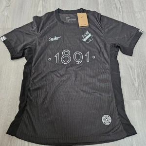 Svart AIK fotbollströja Smokinglirare Edition 2025 - Säljer en svart AIK fotbollströja från Nike, Smokinglirare Edition 2025. Tröjan har korta ärmar, vit AIK-logga och texten 1891 på bröstet. Materialet är lätt och andas, perfekt för match eller träning. Snygga vita detaljer och diskret mönster i tyget.