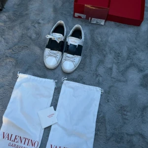 Valentino skor - Säljer ett par vita Valentino Garavani Open sneakers med bred svart rem över vristen och klassiska nitar på sulan. Skorna har rund tå, snörning och platt sula. Kommer med originalpåse och låda.